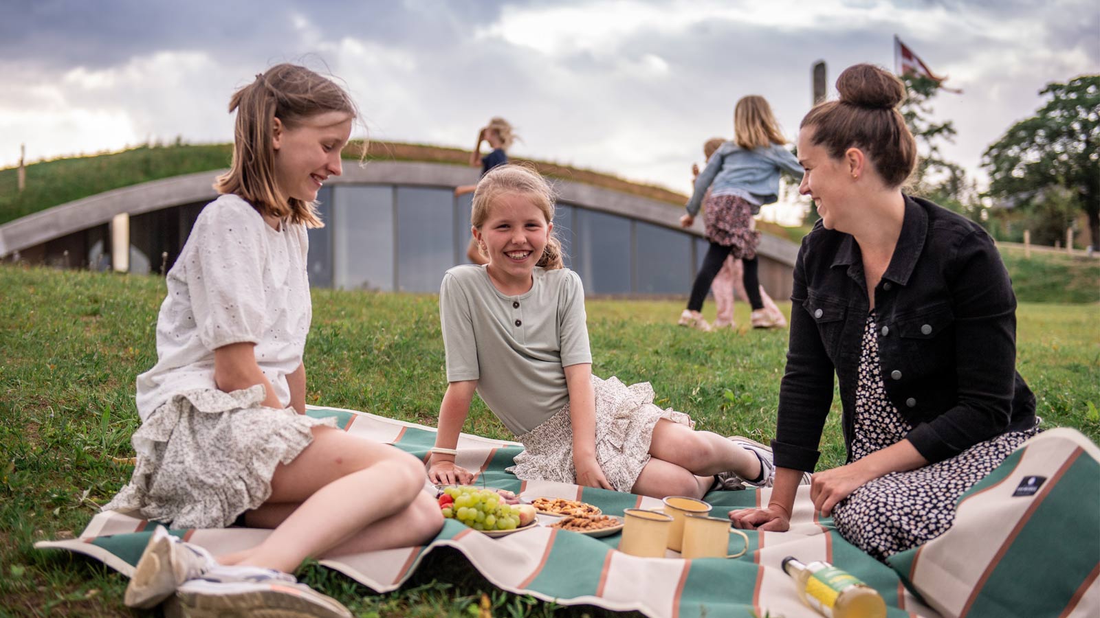 En mor og hendes to døtre sidder på et tæppe og spiser snacks. Bagved ligger besøgscenteret, bygget ind i en jordvold.