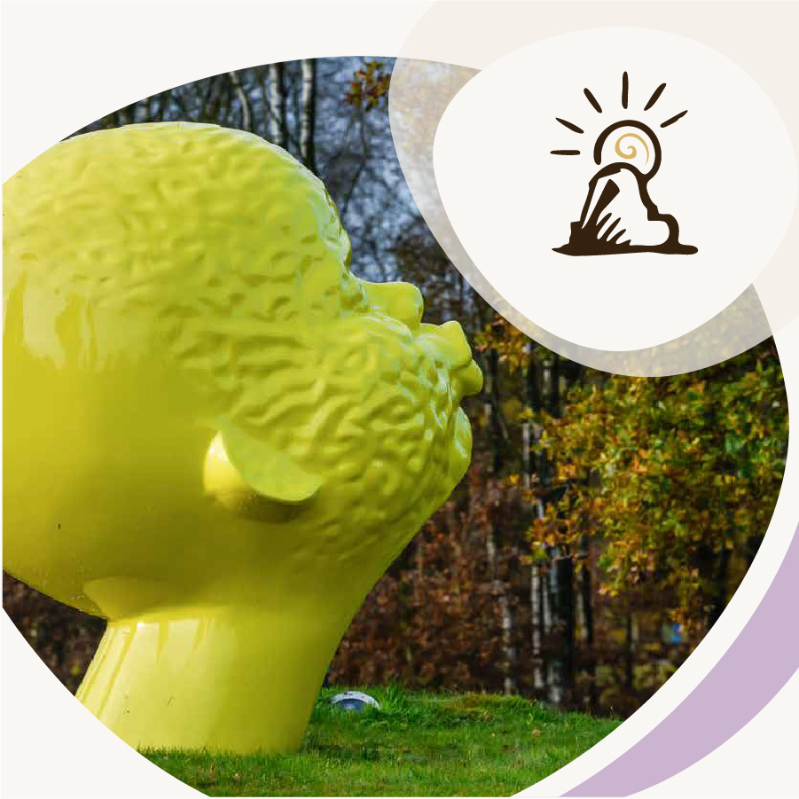 Stor gul kunstskulptur af et hoved i græslandskab, med ikon af en skulptur og sol øverst til højre.