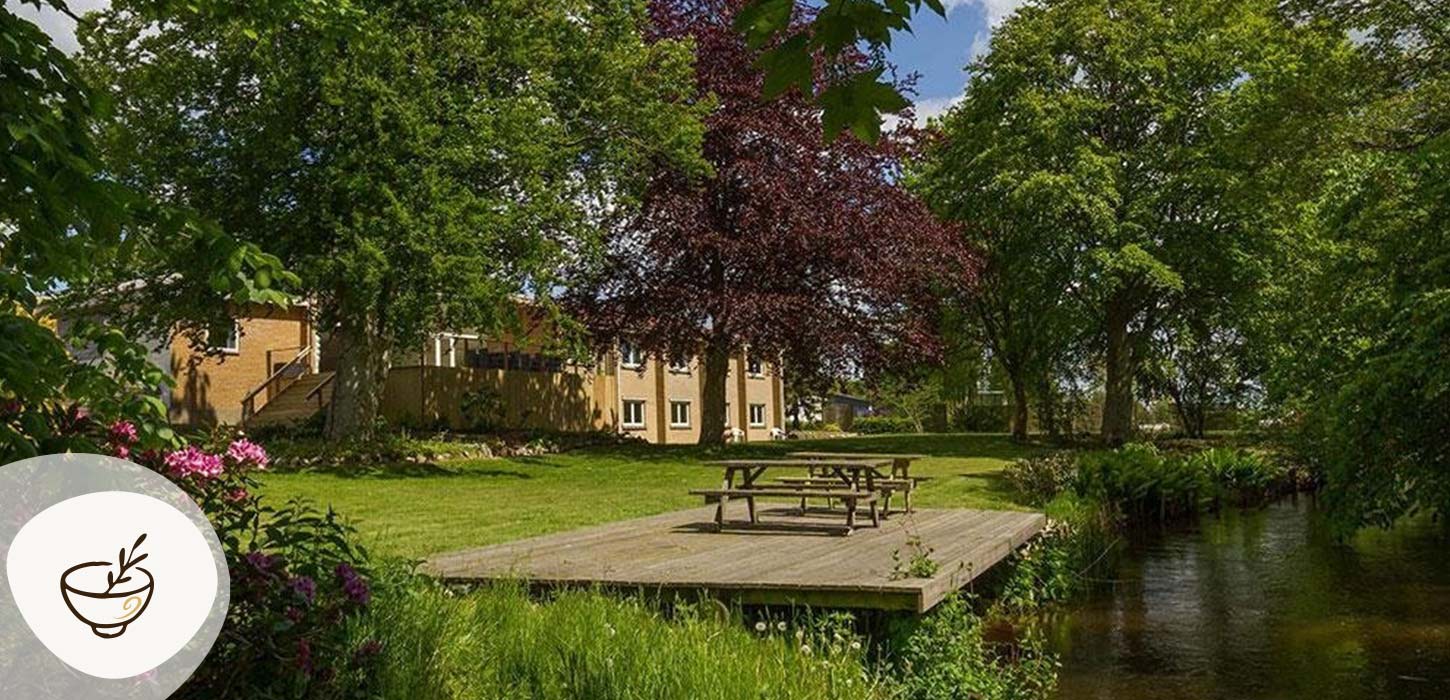 Omme Å, have og terrasse med borde-bænke-sæt bagved Sdr. Omme Kro, omgivet af grønne træer og rolige omgivelser. Kroen er en del af Gastroruten i Small Wonders