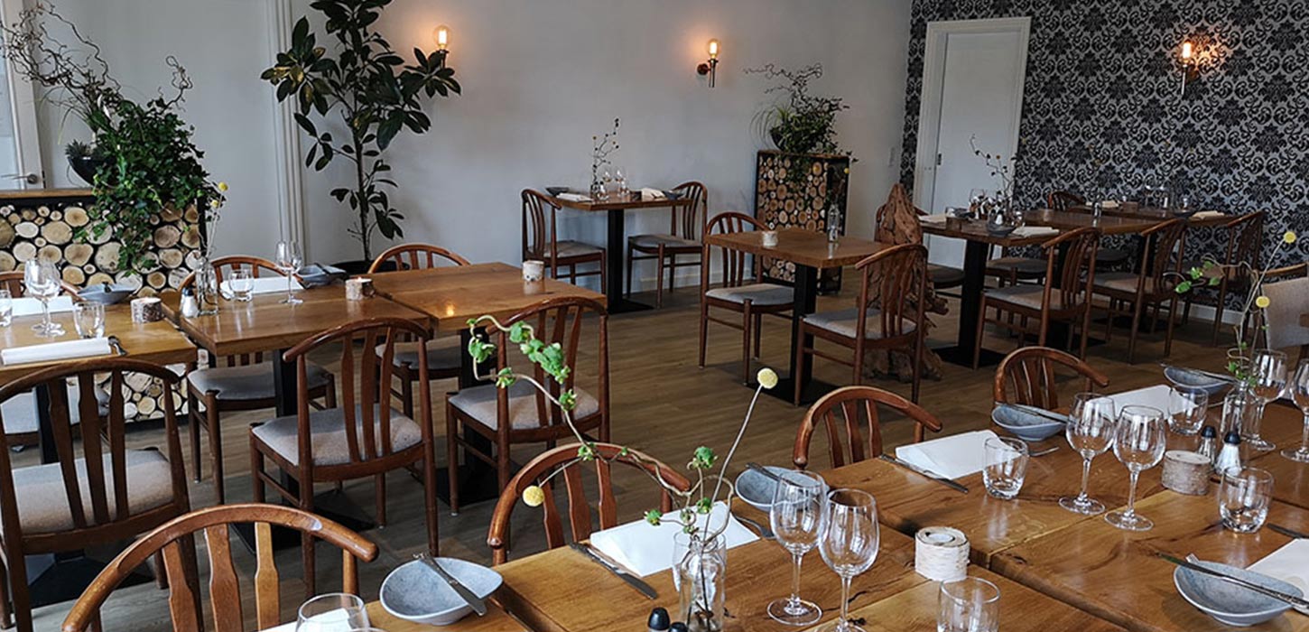 Stemningsfuld spisestue på Sdr. Omme Kro med dækkede træborde, klassiske træstole og grønne dekorationer. En af spiseoplevelserne på Small Wonders-ruterne.