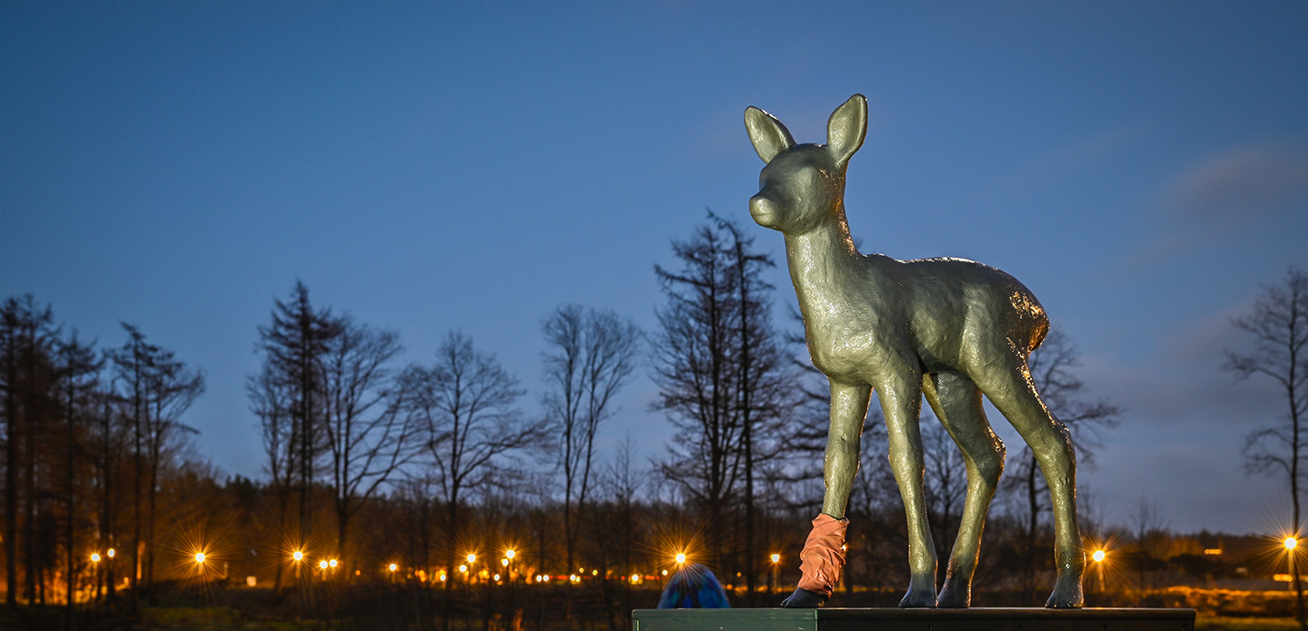 Skulpturen Råbarn i Skulpturpark Billund, vist i aftenlys med træer og parkens lamper i baggrunden. En del af Kunstruten i Small Wonders – Heart of Jutland.