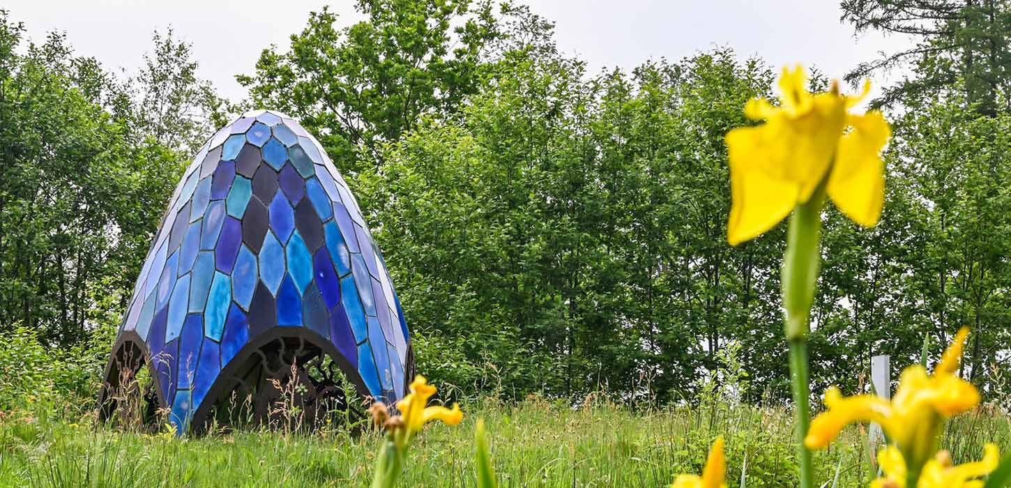 Skulpturen Pentagonia i Skulpturpark Billund, en blå mosaikformet installation med udskårne metaldetaljer ved basen. Gule påskeliljer foran skulpturen. En del af Kunstruten i Small Wonders – Heart of Jutland.