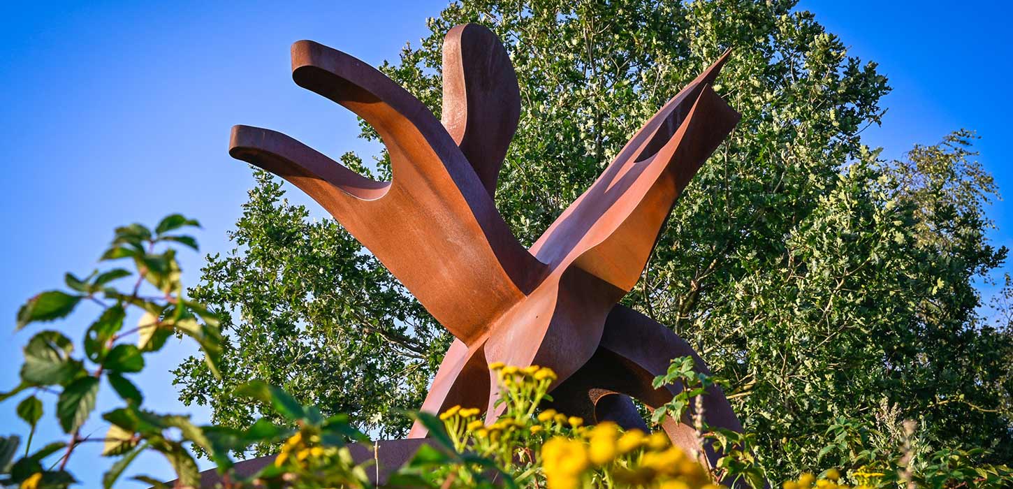 Skulpturen STJERNEDYRET i Skulpturpark Billund, set nedefra mellem grønne planter. og gule blomster. En del af Kunstruten i Small Wonders – Heart of Jutland.